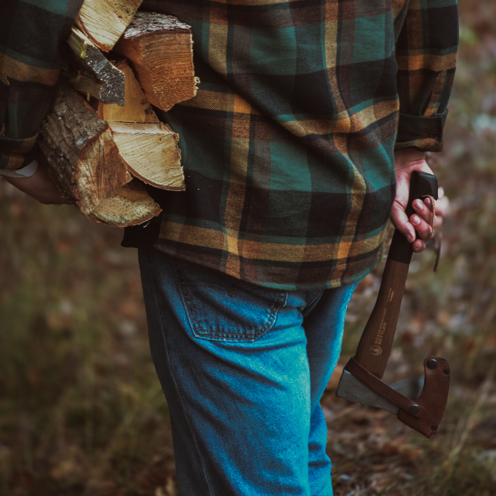 Person med flanellskjorta och yxa som bär ved i skogsmiljö