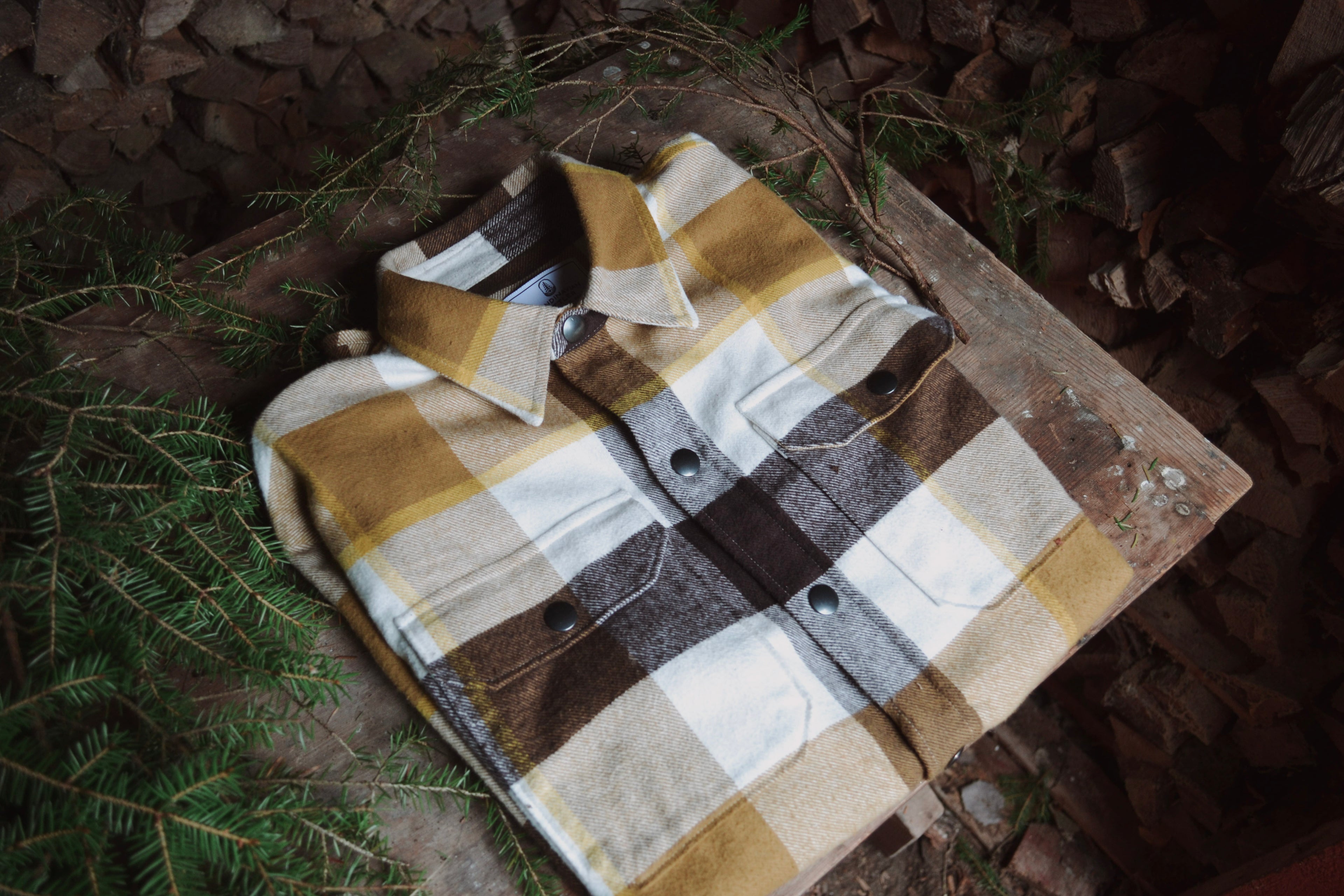 Hopvikt rutig flanell-overshirt i brunt och beige liggande bland barrgrenar.